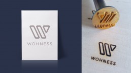 Logodesign der Firma Wohness und daneben die Anwendung des Logos mit einem Brandstempel in Holz gebrannt